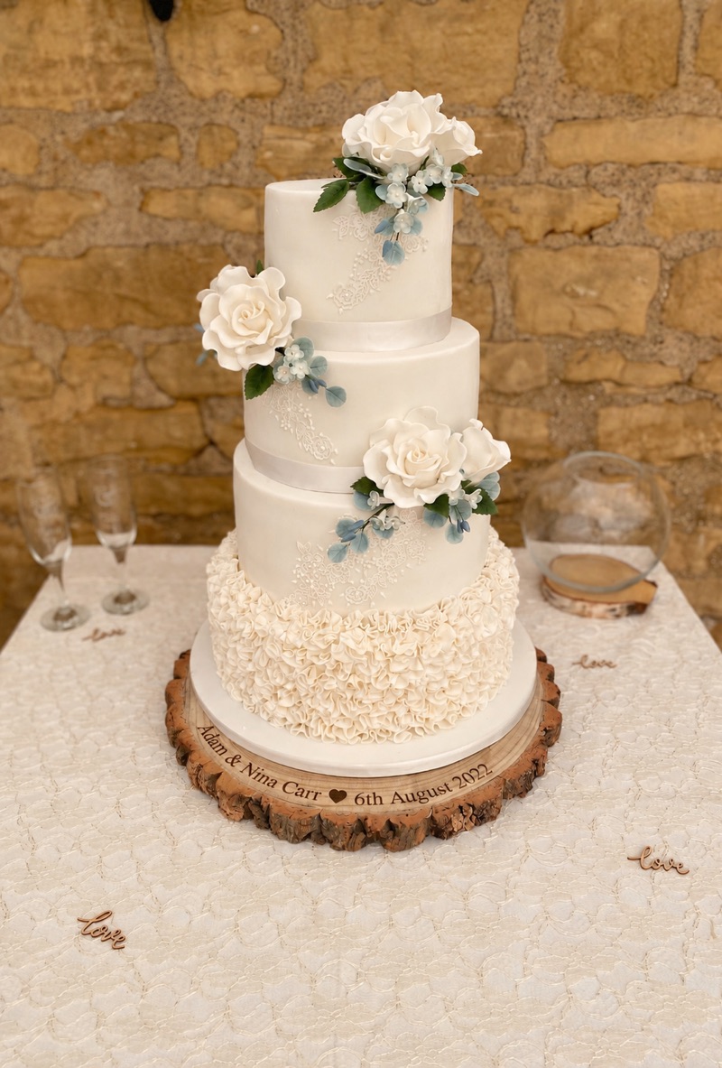Four-tier ivory wedding cake with white roses, lace details and a ruffled lower tier