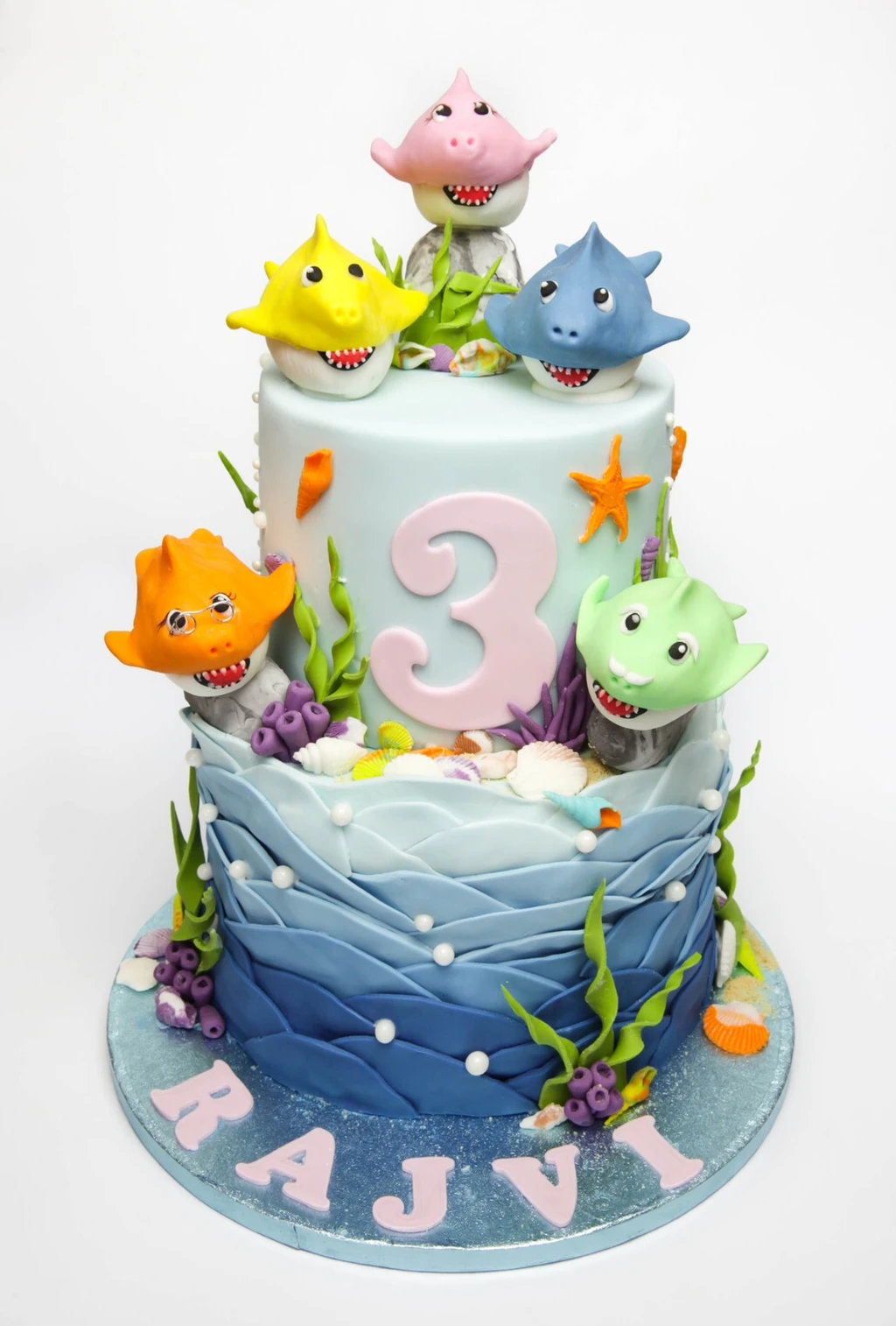 Two-tier ocean-themed third birthday cake with colourful shark figures, shells and blue waves