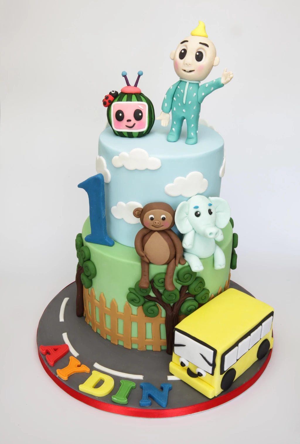 Two-tier first birthday cake with a baby figure, cartoon watermelon, animals and a yellow bus